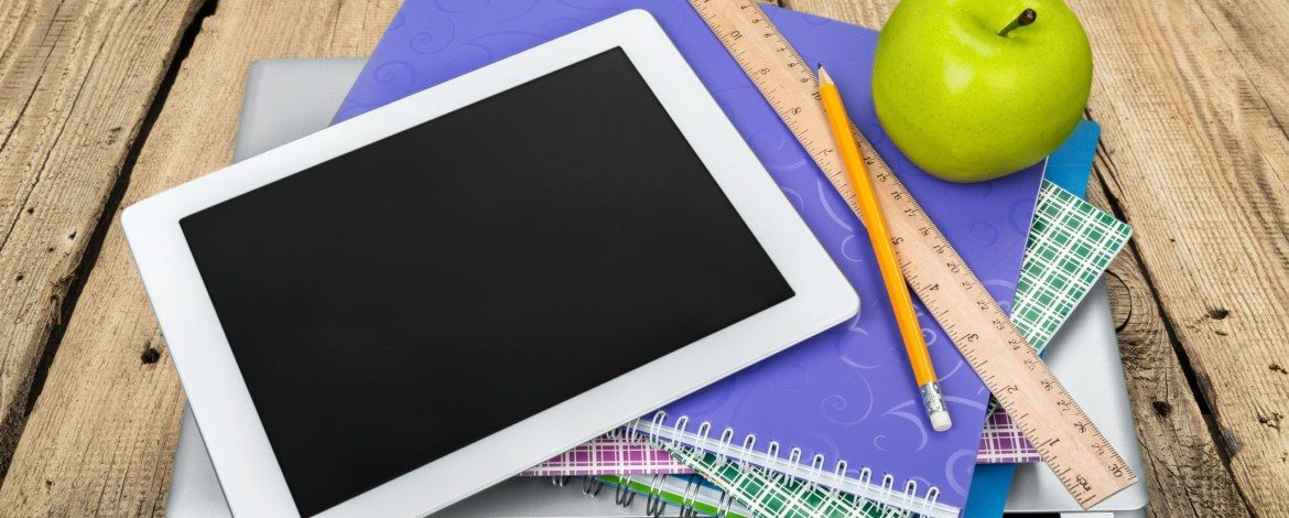 A white iPad on a pile of school copy books. There is also a pencil, a ruler and a green apple there.