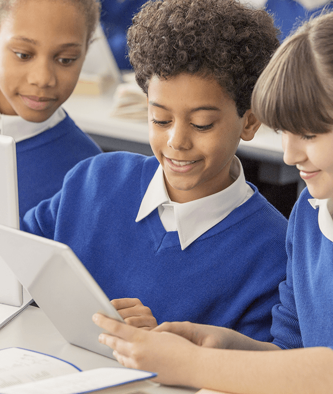 school pupils in blue jumper are at their desk looking at an iPad. They are smiling as they touch the screen.